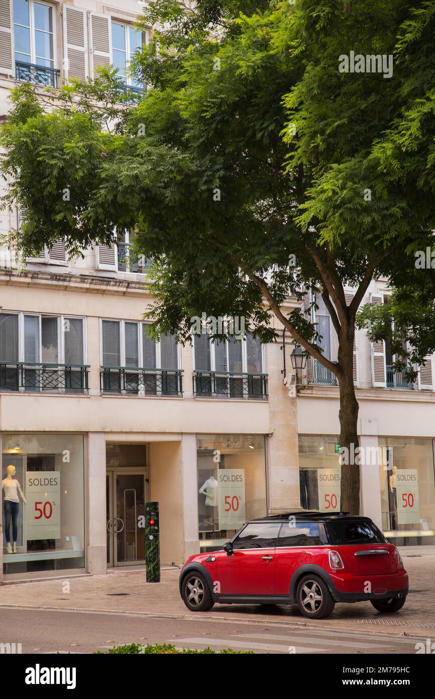 In der Stadt steht ein kleines rotes Auto unter einem grünen Baum. Das Auto steht in der Nähe einer Fußgängerüberquerung vor einem Gebäude mit Gewerbeflächen Stockfoto In der Stadt steht ein kleines rotes Auto unter einem grünen Baum. Das Auto steht in der Nähe einer Fußgängerüberquerung vor einem Gebäude mit Gewerbeflächen Stockfoto