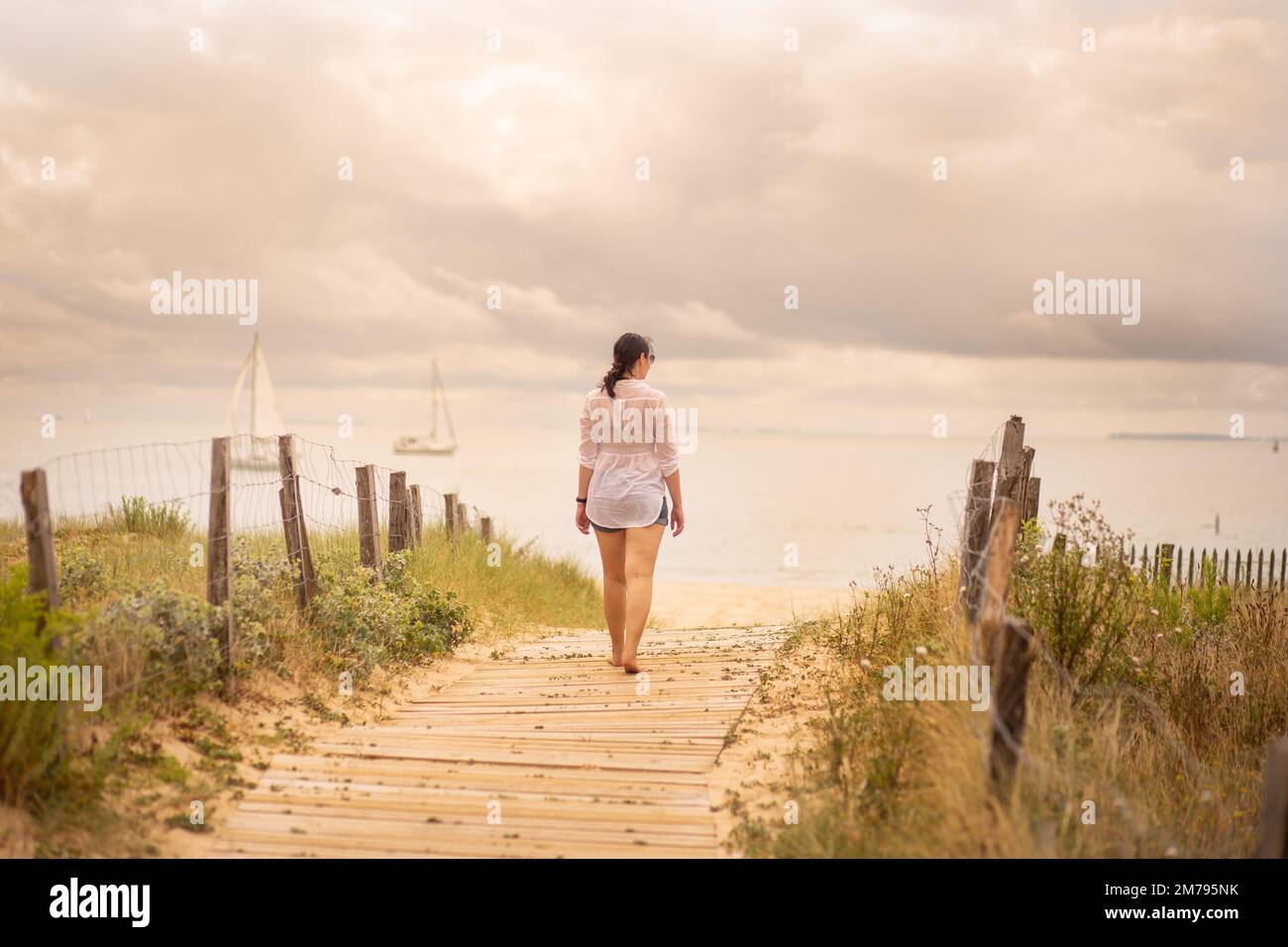 Eine junge Frau geht auf dem hölzernen Ponton, der zum Strand führt. Im Hintergrund können Sie zwei Boote mit weißen Segeltörn sehen, die im ruhigen Wasser segeln Stockfoto