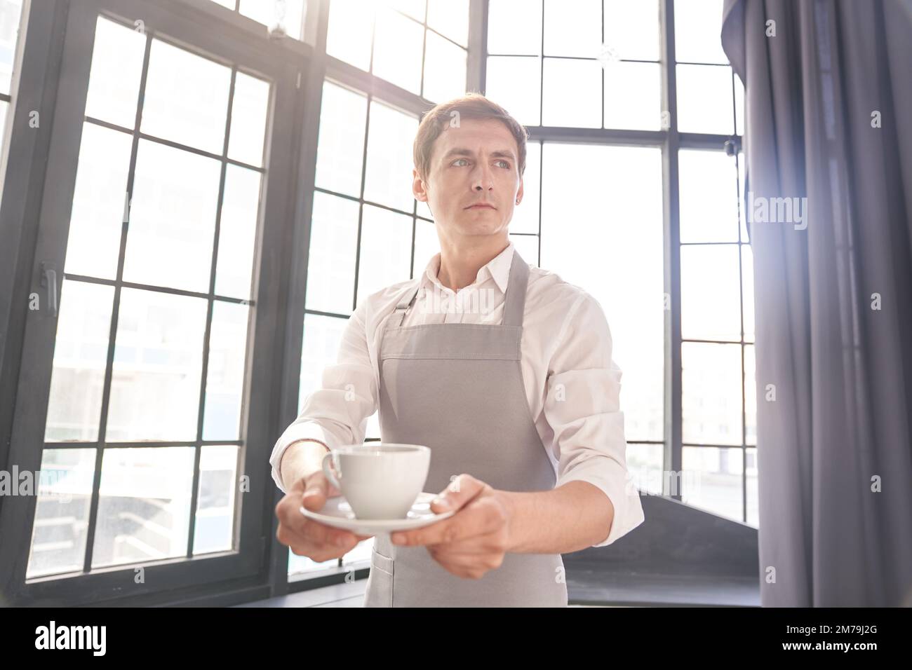 Ein Kellner in einer grauen Schürze hält eine Tasse Kaffee. Der Barista gibt eine Tasse heißen Kaffee in einem Café vor dem Hintergrund eines großen Fensters. Hochwertiges Foto Stockfoto