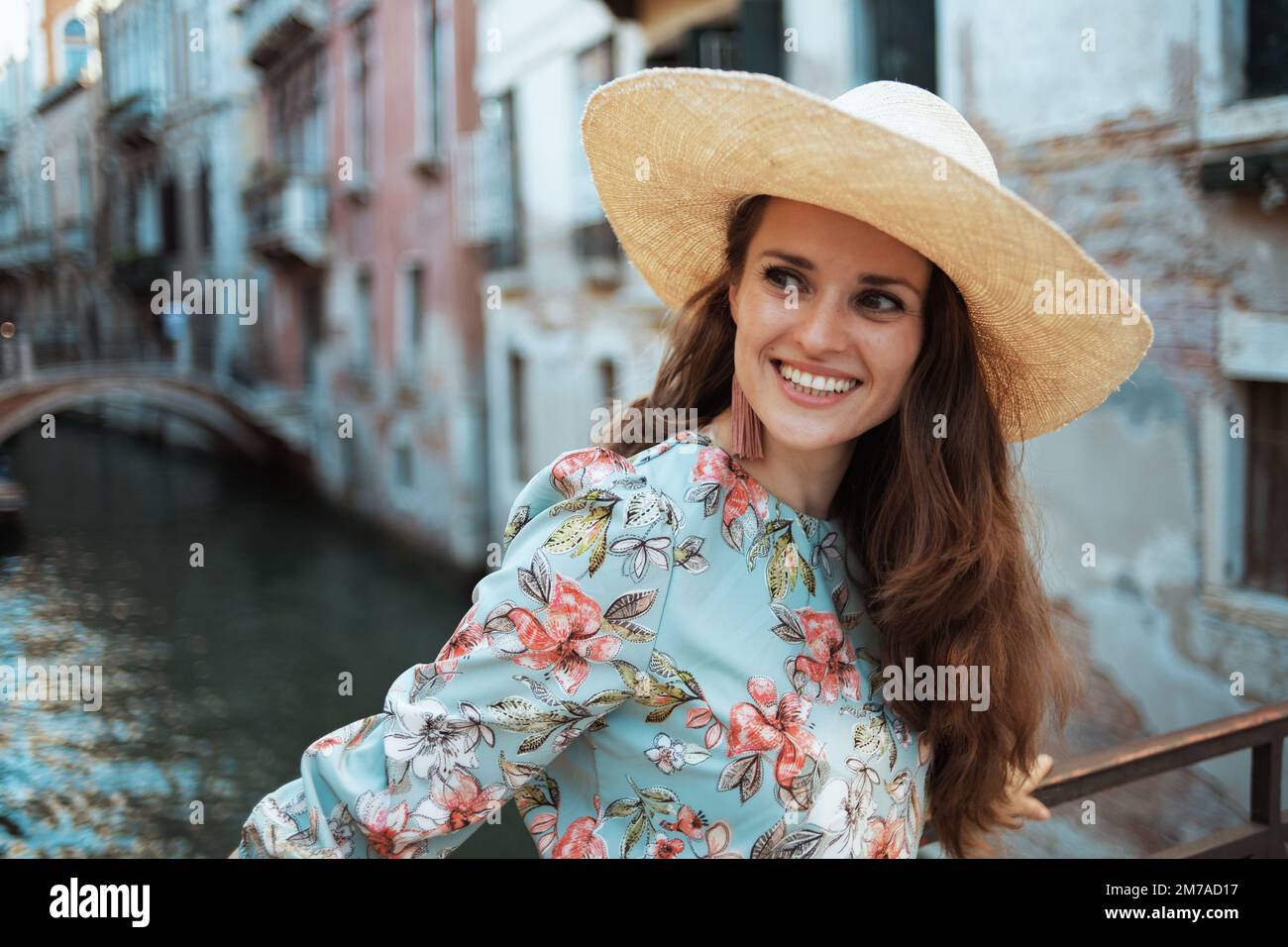 Eine glückliche, moderne Alleinreisende in Blumenkleid mit Hut genießt die Promenade in Venedig, Italien. Stockfoto