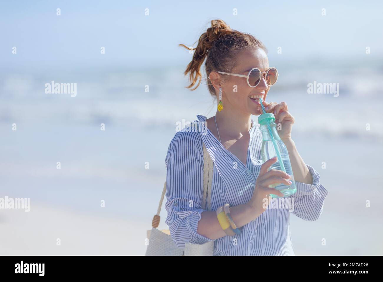 Lächelnde elegante 40-jährige Frau mit weißem Strohbeutel und Getränk am Strand. Stockfoto