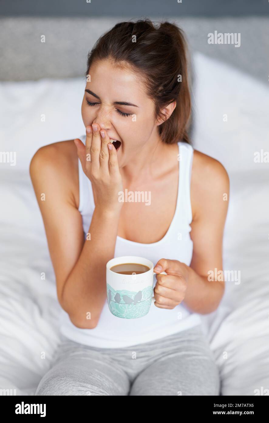 Immer noch so schläfrig... Eine junge Frau gähnt, während sie mit einer Tasse Kaffee im Bett sitzt. Stockfoto