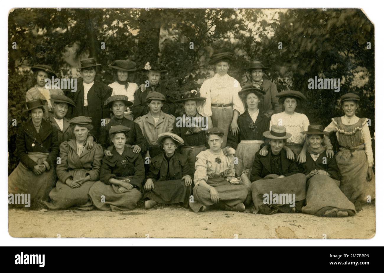 Originelle Postkarte aus der späten Edwardianischen Ära mit einer Gruppe von Arbeiterfrauen, viele Figuren unterschiedlichen Alters. Viele tragen die Damenversion der flachen Mütze. Es wird angenommen, dass es Hopfenpflücker sind, wie es in einer ländlichen Umgebung zu sein scheint, ist eine große Gruppe von verschiedenen Altersgruppen und einige tragen Positionen, die wie ein Haufen Hopfen am Revers aussehen, und tragen Schürzen, die an der Taille gebunden sind, über urbaner Kleidung. Viele Londoner Arbeiter haben während eines Arbeitsurlaubs in den Kent Hop Fields gearbeitet. Ungefähr 1905. Stockfoto