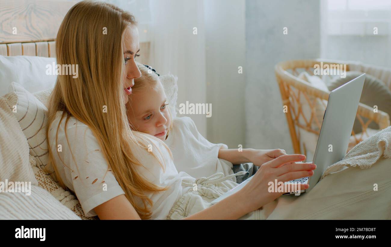 Kaukasische Mutter mit Tochter, die ihren Laptop im Online-Shop im Bett surft, im Internet einkauft und zusammen im Schlafzimmer einen Film anschaut. Mom unterrichtet Little Stockfoto