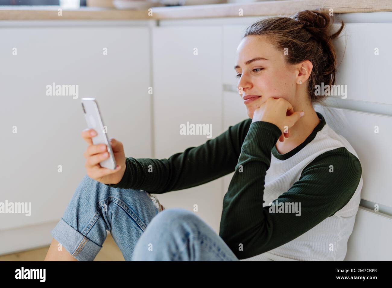 Junge Frau sitzt mit Smartphone und scrollt in einer Küche. Stockfoto