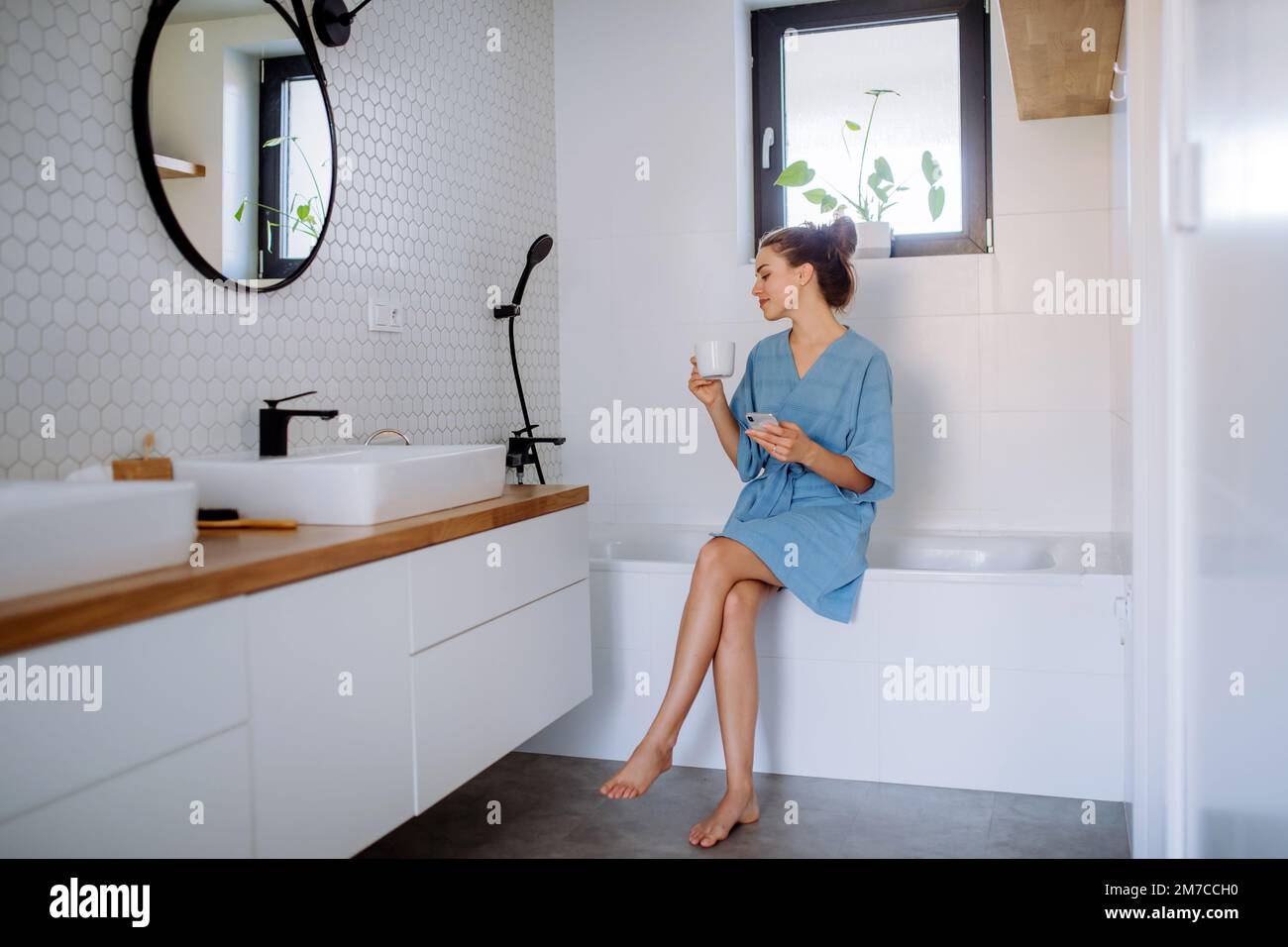 Junge Frau im Badezimmer mit Telefon und Tasse Kaffee, Morgenroutine und Entspannungskonzept. Stockfoto