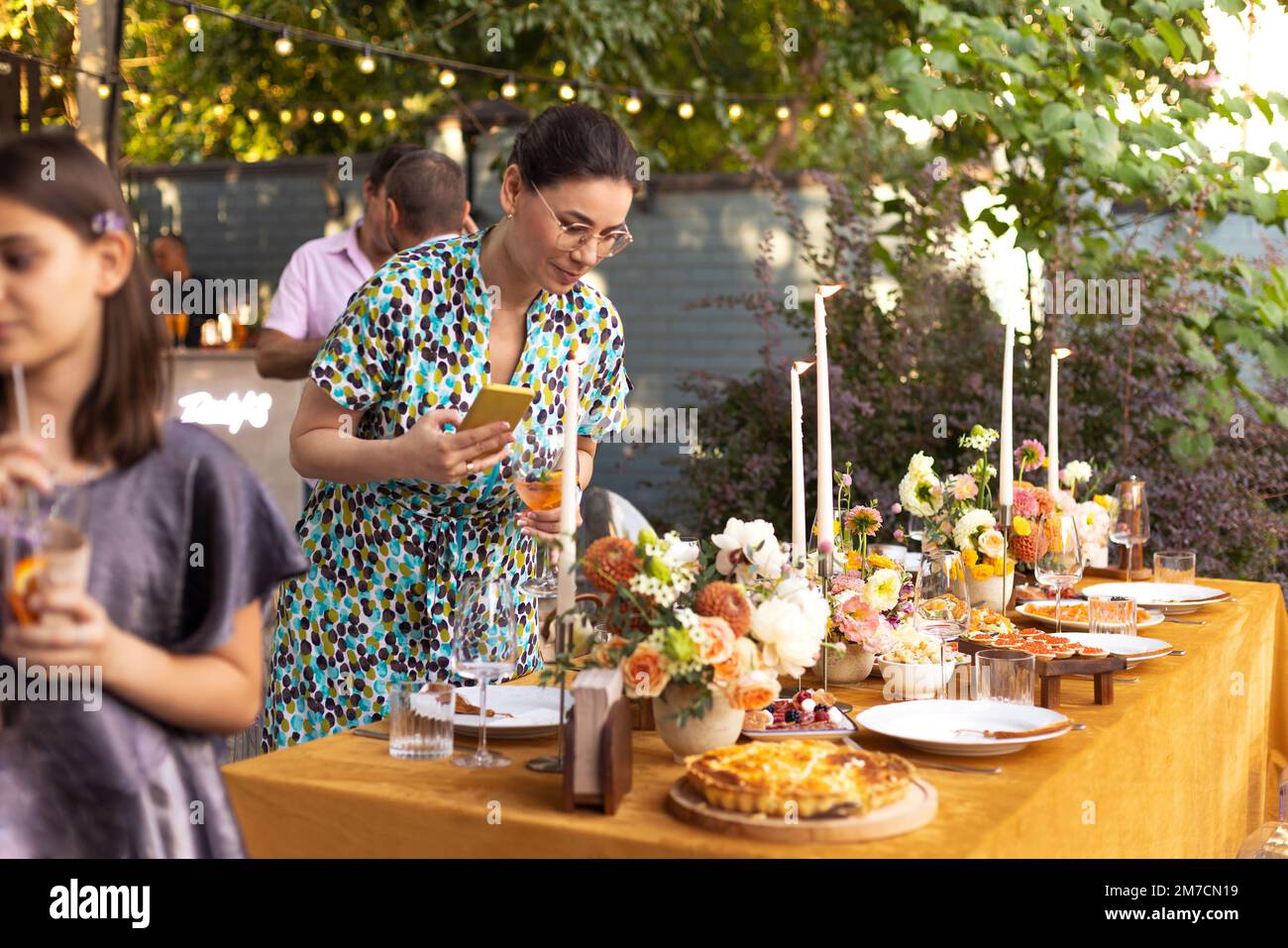 Eine herzliche, brünette Frau hält ein Telefon und macht ein Foto von einem Hochzeitstisch, ein Foto von einer Hochzeitsdekoration für ein 20-jähriges Jubiläum, eine Gartenparty Stockfoto