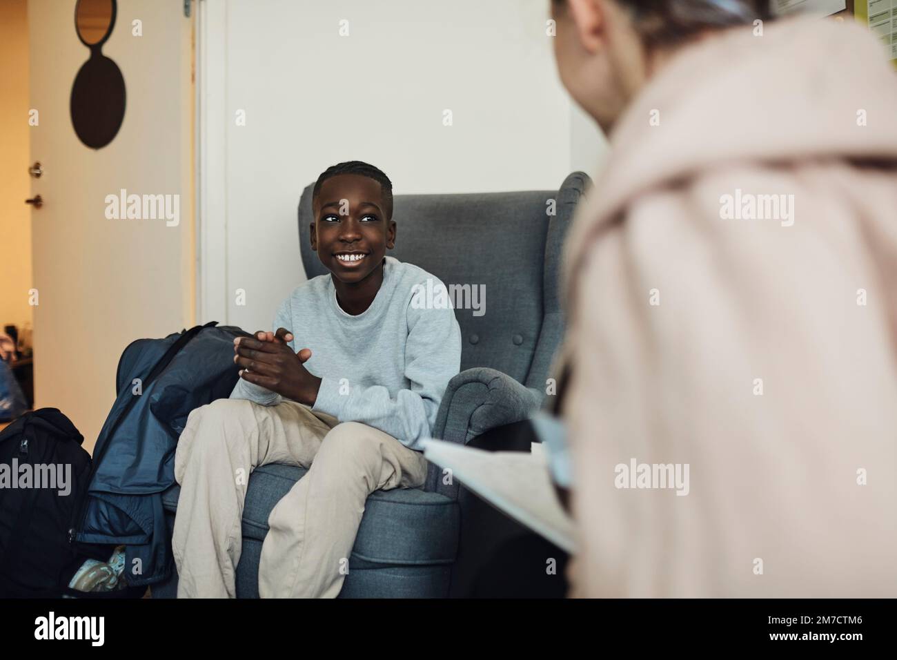 Lächelnder Teenager, der mit einem Psychiater spricht, während er in der Schulpraxis auf einem Stuhl sitzt Stockfoto