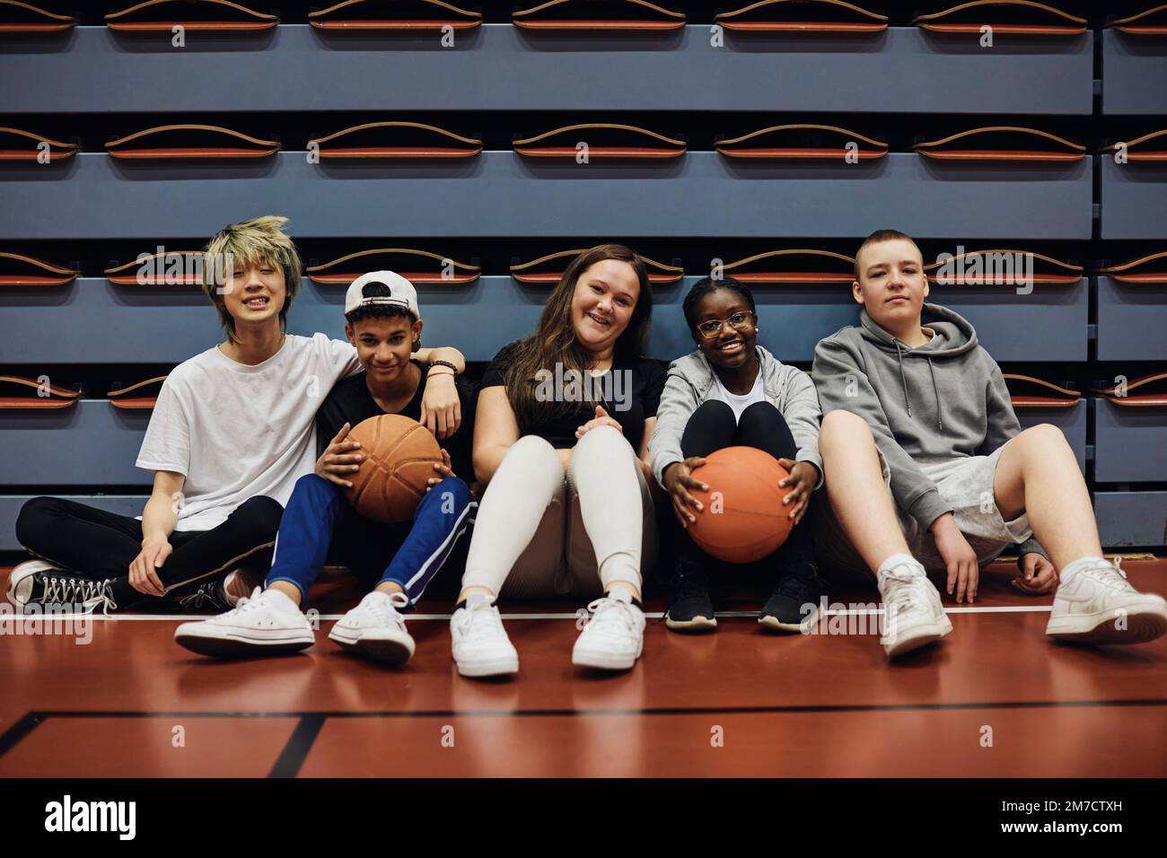 Porträt lächelnder männlicher und weiblicher Freunde, die mit Basketbällen auf dem Sportplatz sitzen Stockfoto