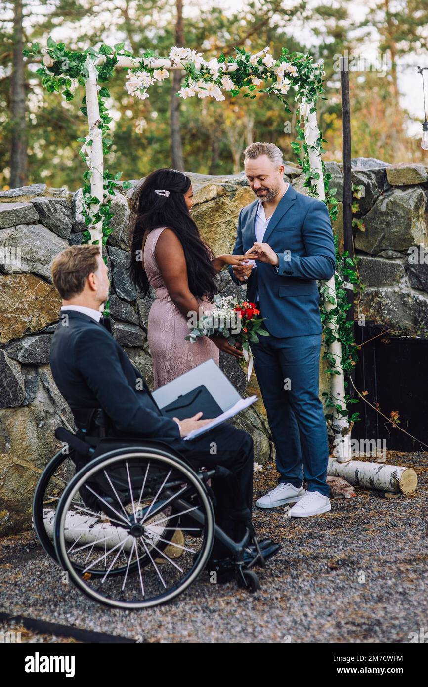 Lächelnder Bräutigam und Braut, Tauschring eines Pfarrers im Rollstuhl mit Behinderung bei der Hochzeitszeremonie Stockfoto