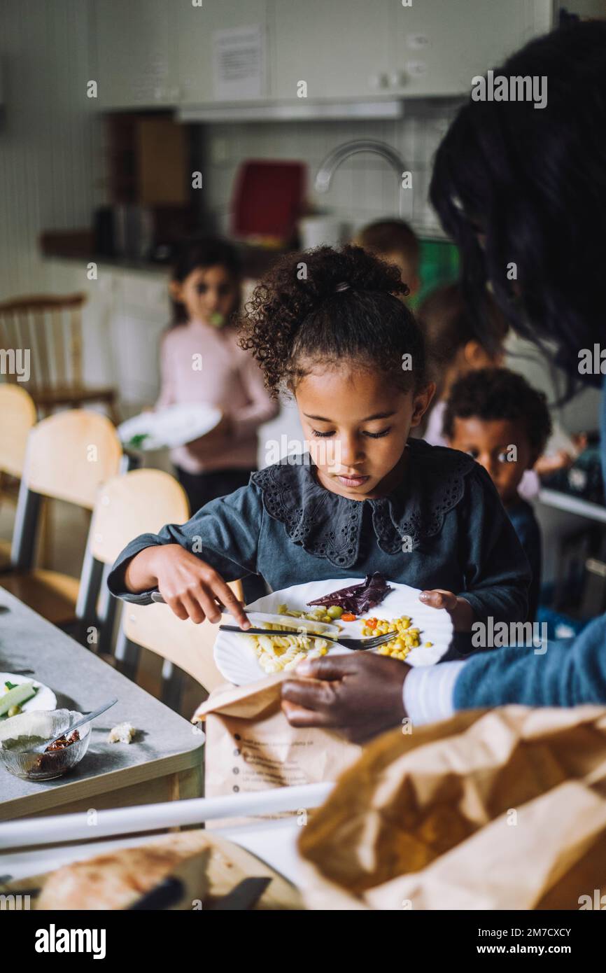 Mädchen, das Essensreste in eine Papiertüte wirft, die der Lehrer in der Kinderbetreuung hält Stockfoto