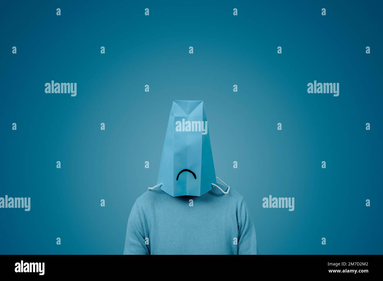 Ein Mann trägt eine blaue Papiertüte in seinem Kopf, mit einem traurigen Mund darin, vor einem blauen Hintergrund mit einem leeren Raum um ihn herum Stockfoto