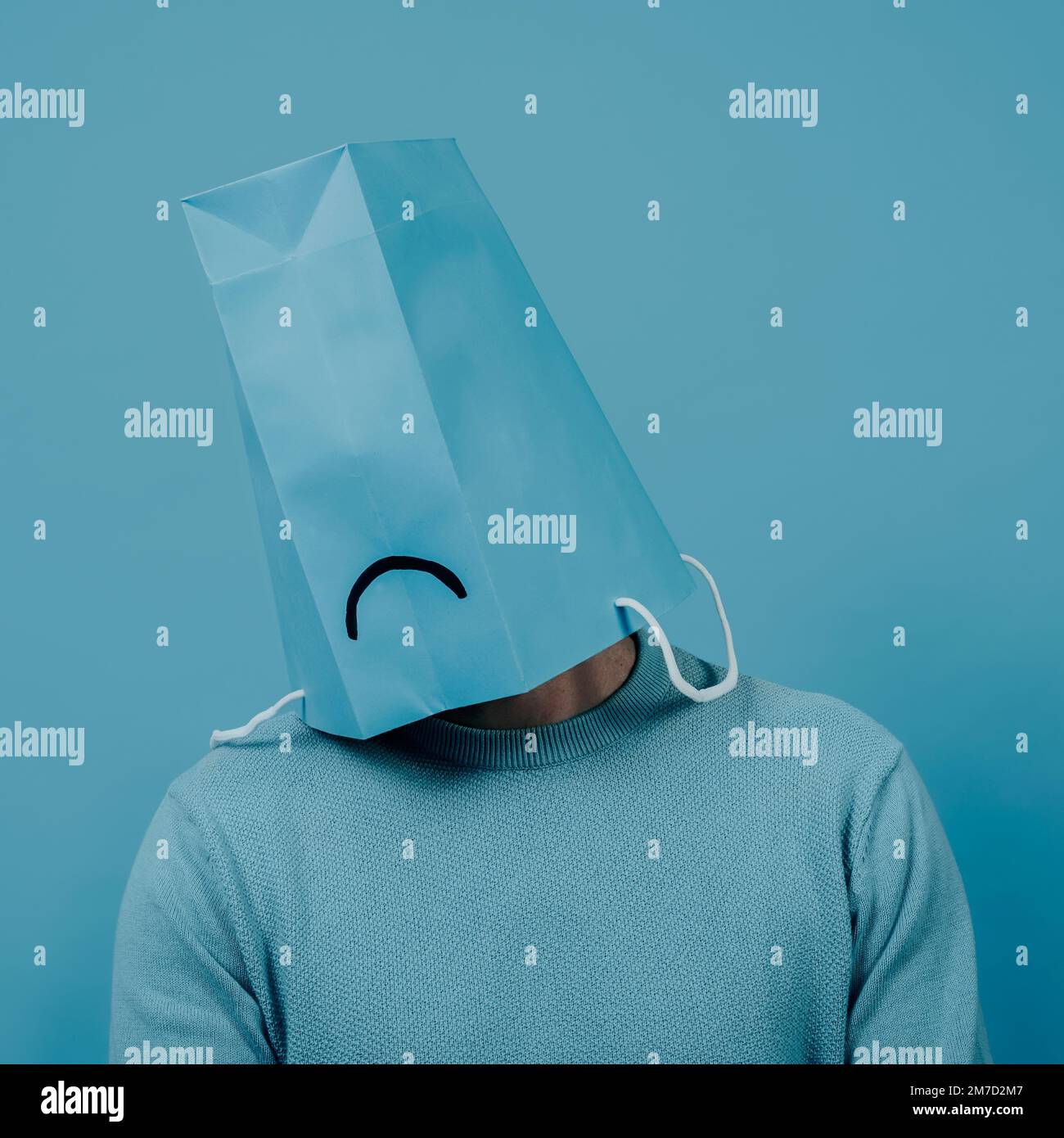 Nahaufnahme eines Mannes, der eine blaue Papiertüte im Kopf trägt, mit einem traurigen Mund darin, vor einem blauen Hintergrund Stockfoto