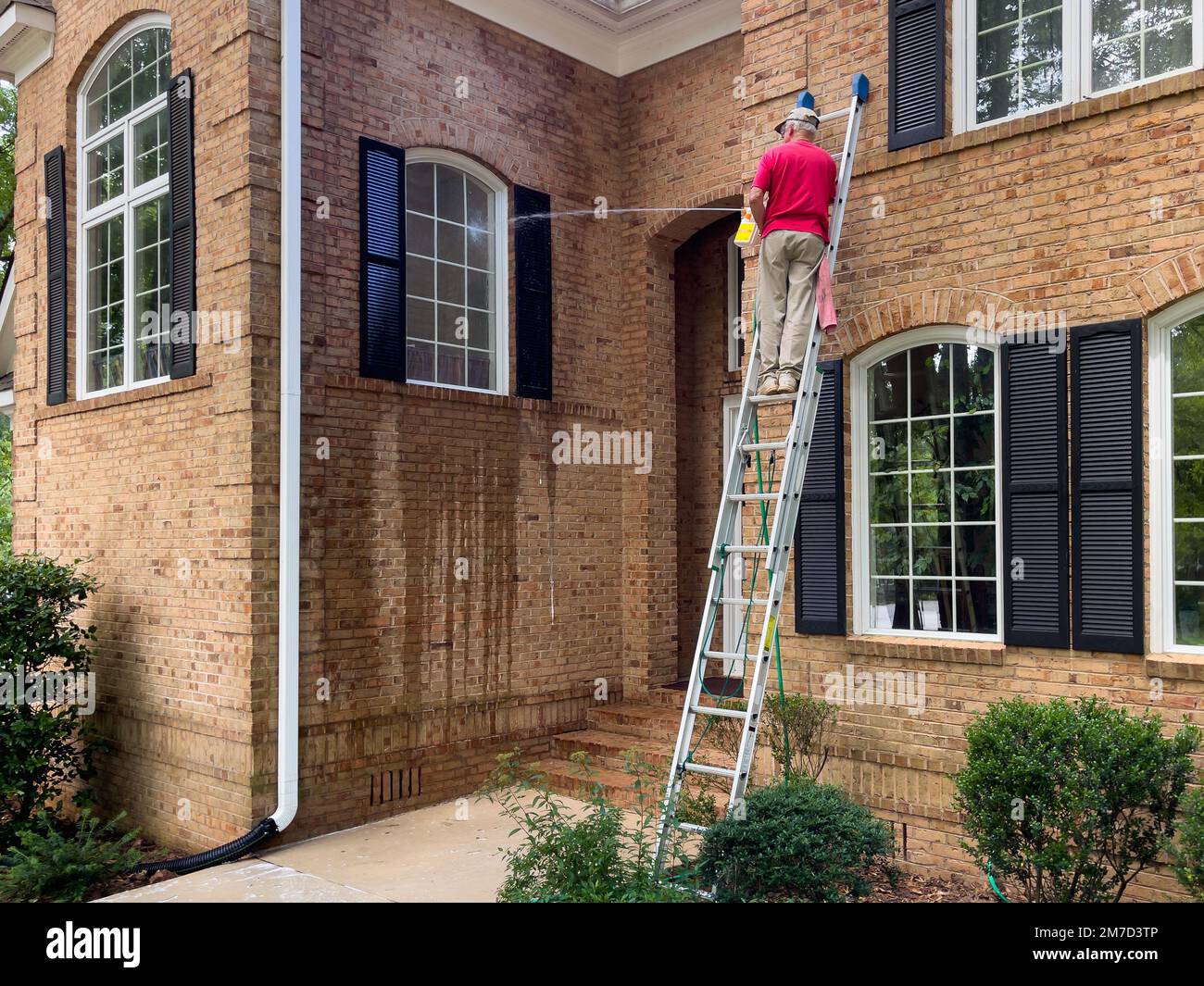 Ein Mann auf einer Leiter wäscht die Fenster eines Hauses mit einem Seifensprühgerät. Federreinigung mit Schlauch und Hochdruckreiniger. Reinigungsservice. Stockfoto