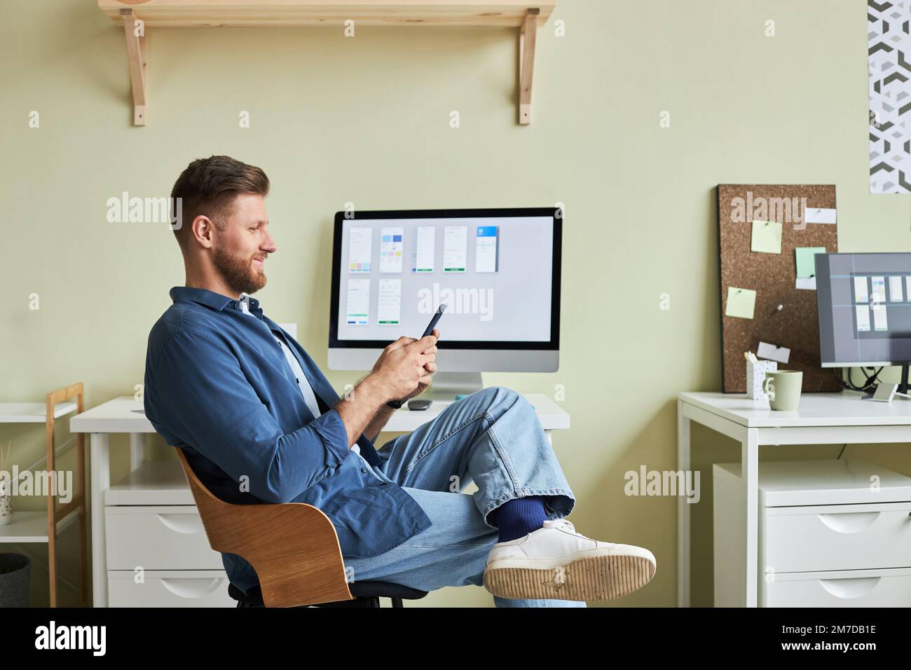 Seitliches Porträt eines lächelnden jungen Mannes, der sich am Arbeitsplatz entspannt und sein Smartphone in minimaler Büroumgebung verwendet Stockfoto