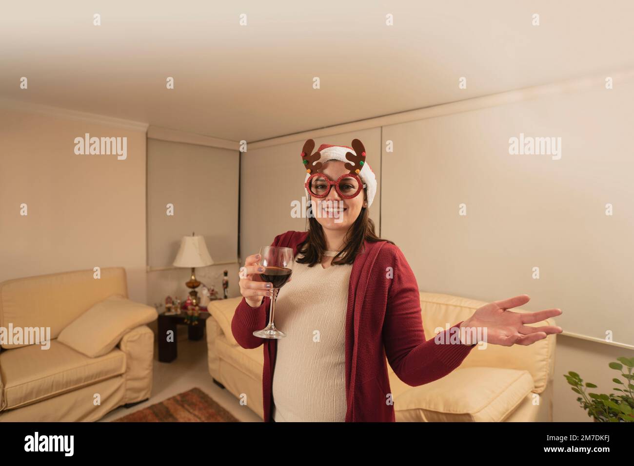 Eine wunderschöne junge hispanikerin, verkleidet mit Rentiergläsern und einem weihnachtshut, die in die Kamera schaut und mit einem Glas Rotwein anstoßen Stockfoto