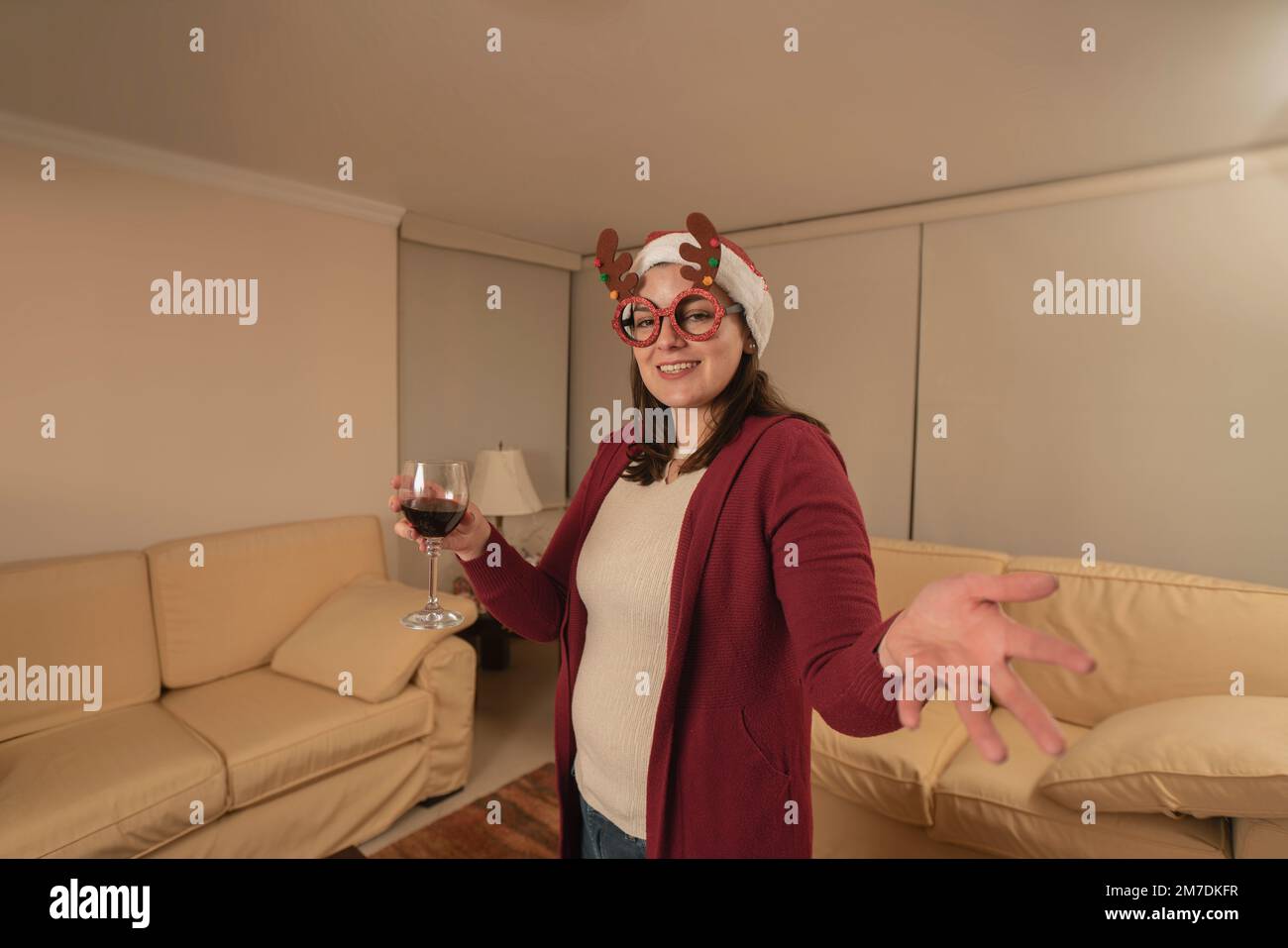 Eine wunderschöne junge hispanikerin, verkleidet mit Rentiergläsern und einem weihnachtshut, die in die Kamera schaut und mit einem Glas Rotwein anstoßen Stockfoto