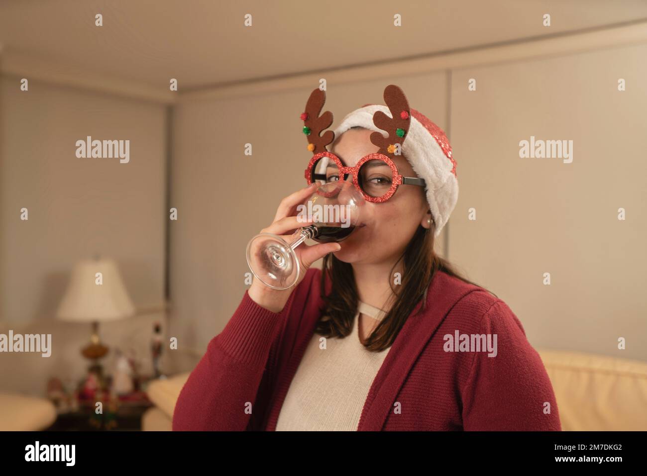 Wunderschöne hispanische junge Frau, verkleidet mit Rentiergläsern und weihnachtsmütze, die nachts in ihrem Wohnzimmer ein Glas Rotwein trank Stockfoto