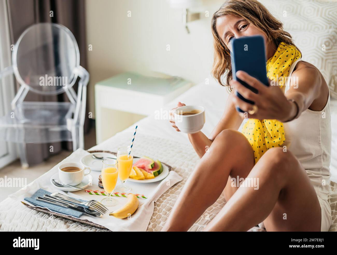 Eine weiße Frau mittleren Alters führt einen Videoanruf, während sie ein Obstfrühstück auf dem Bett in einem Hotelzimmer isst. Sie hält einen Kaffee in der Hand. Stockfoto