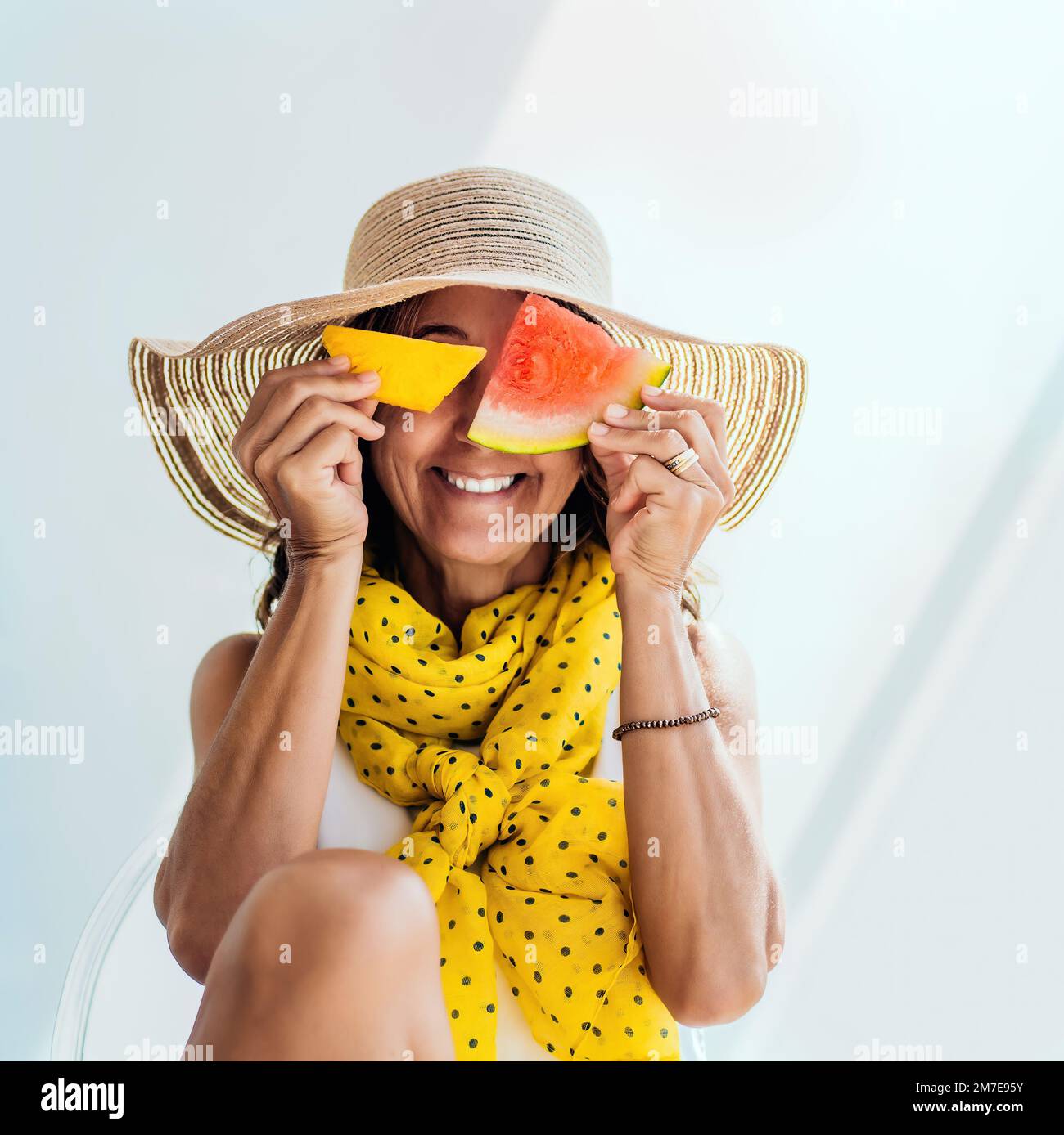 Eine Frau mittleren Alters mit Strohhut und gelbem Schal macht Witze und amüsiert sich, indem sie ihr Gesicht mit einem Stück Ananas und einer Wassermelone bedeckt Stockfoto
