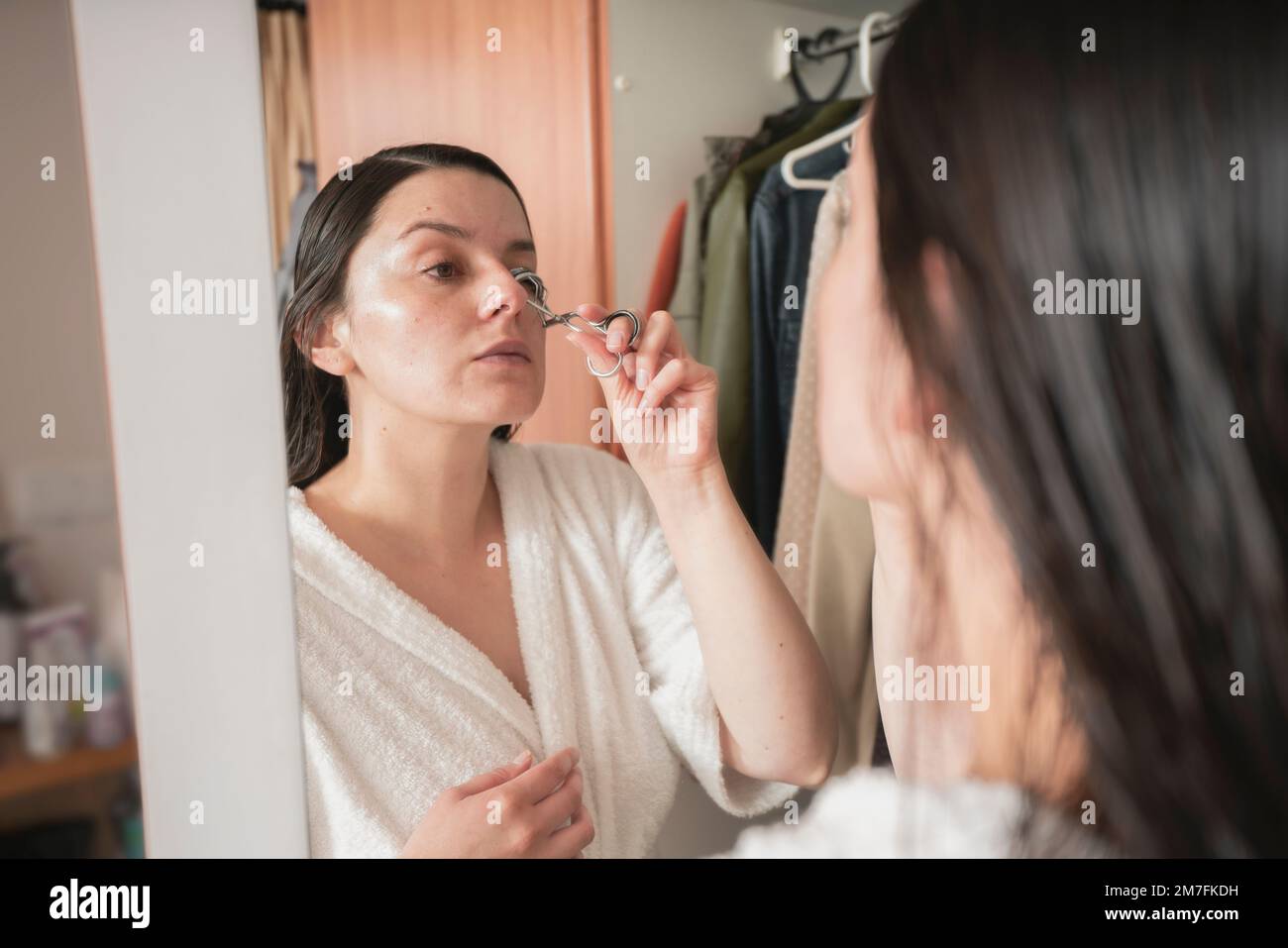 Wunderschöne lateinamerikanische Frau in einem weißen Bademantel, die ihre Augenbrauen vor dem Spiegel an ihrer Schranktür in ihrem Schlafzimmer kämmt Stockfoto