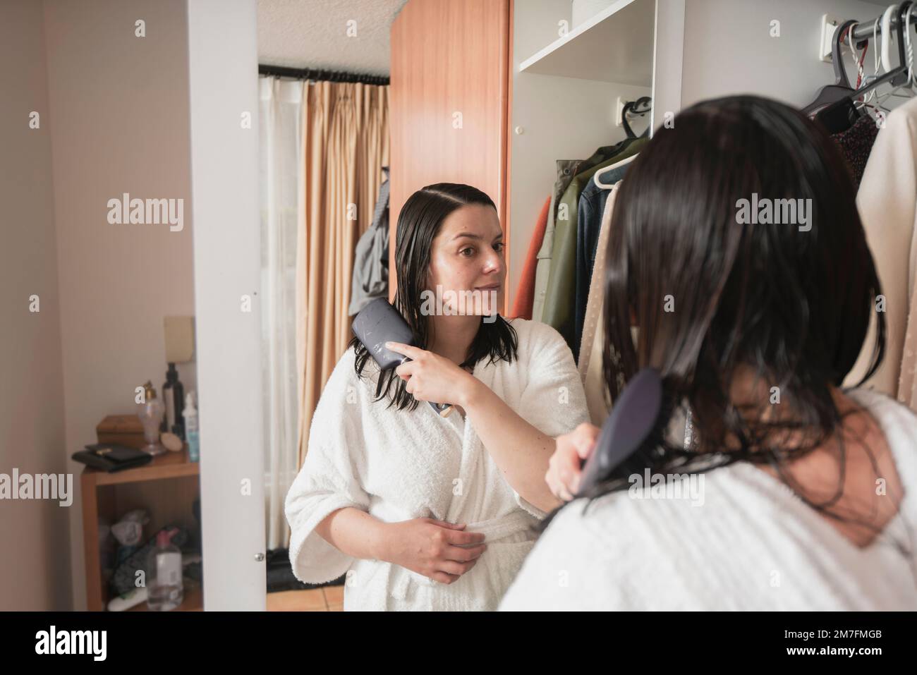 Wunderschöne hispanische Frau in einem weißen Bademantel, die ihr Haar mit einer Bürste vor dem Spiegel kämmt, an ihrer Schranktür in ihrem Schlafzimmer Stockfoto