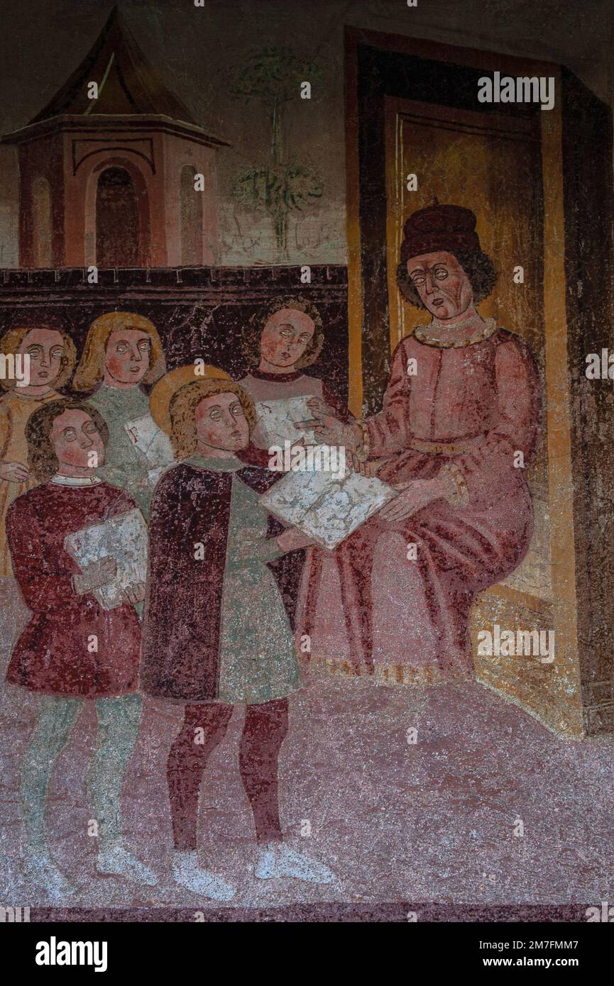 Ein Schuljunge und zukünftiger christlicher heiliger, Anthony der Abt oder Anthony der große, stellt seine Schularbeit einem sitzenden Lehrer vor. Fresko aus der Vorrenaissance des späten 15. Jahrhunderts von einem Mitglied der lombardischen Baschenis-Familie an der Außenmauer der Chiesa di Sant’Antonio Abate (Kirche des Heiligen Antonius des Abtes) in Pelugo, Trentino-Südtirol, Italien. Teil eines Freskenzyklus, der die Lebensgeschichte des heiligen Antonius des Abtes und ein Beispiel einer biblia pauperum erzählt. Stockfoto