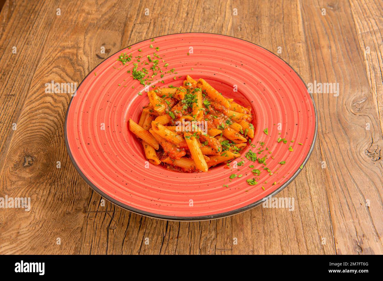 Schöner roter Porzellanteller mit Penne Rezept mit Tomaten und Petersilie auf einem Holztisch Stockfoto