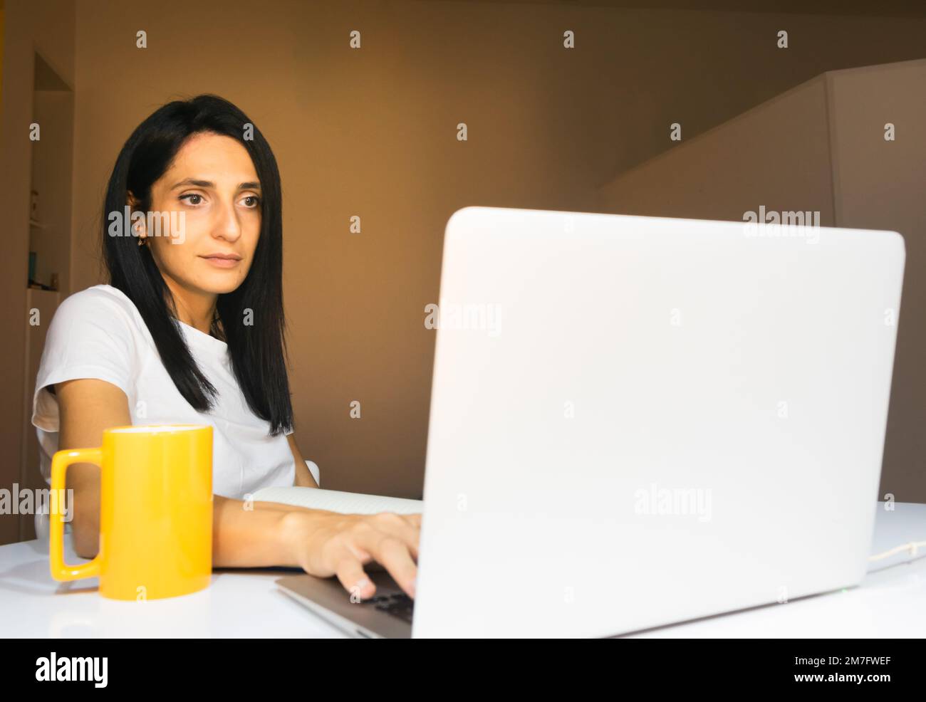 Die junge kaukasische Frau sitzt in der Wohnung hinter dem weißen Schreibtisch und schaut geradeaus. Konzept der Arbeit von zu Hause aus. Stockfoto