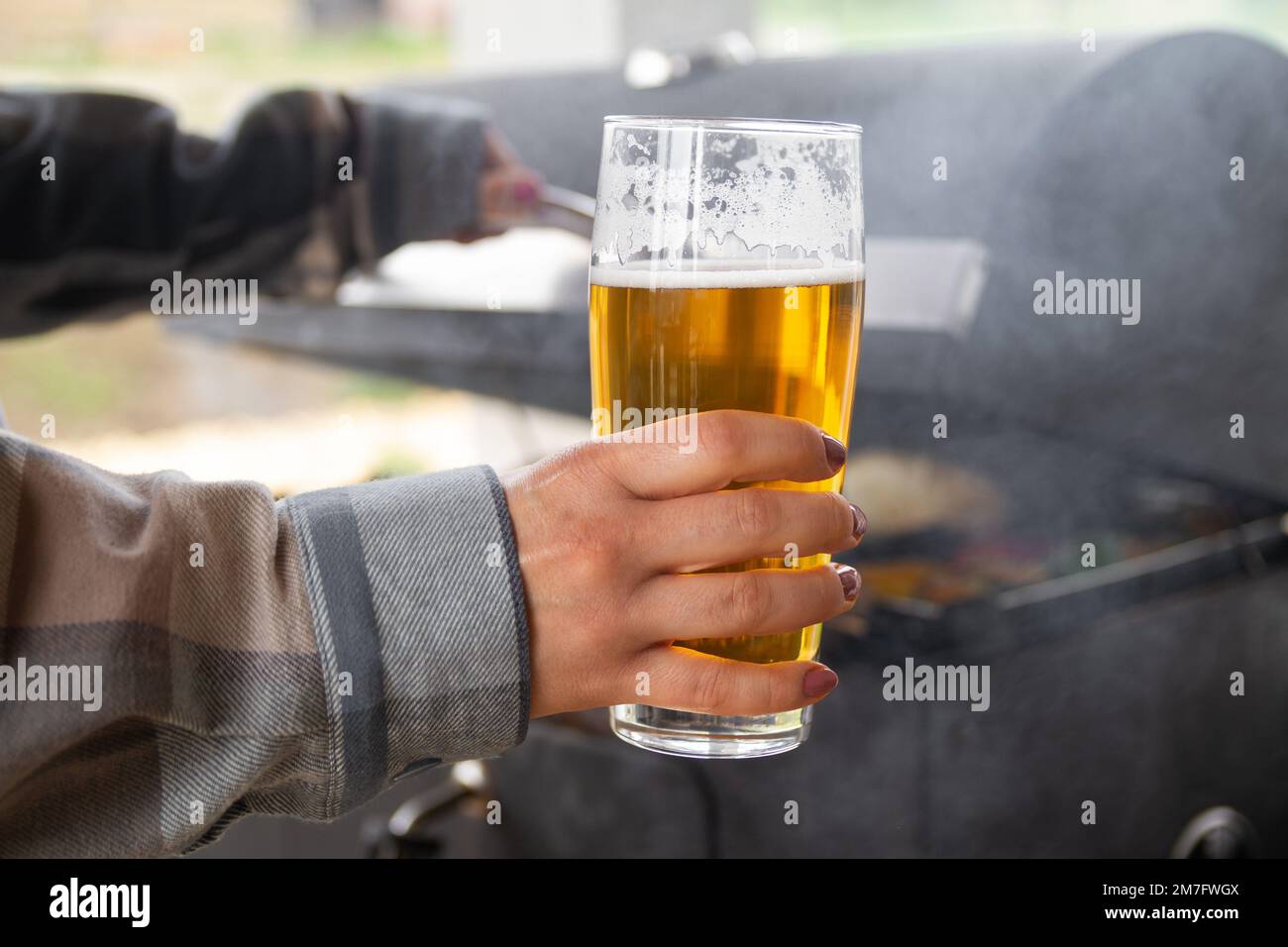 Weibliche Hand hält ein Glas Bier, während sie auf einem Grill kocht. Stockfoto