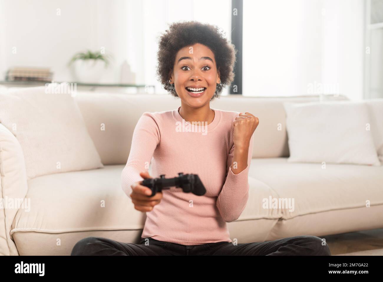 Fröhlich aufgeregte junge gemischte Rassenfrau mit Joystick, die Computerspiel spielt, macht Sieg und Erfolgsgeste Stockfoto