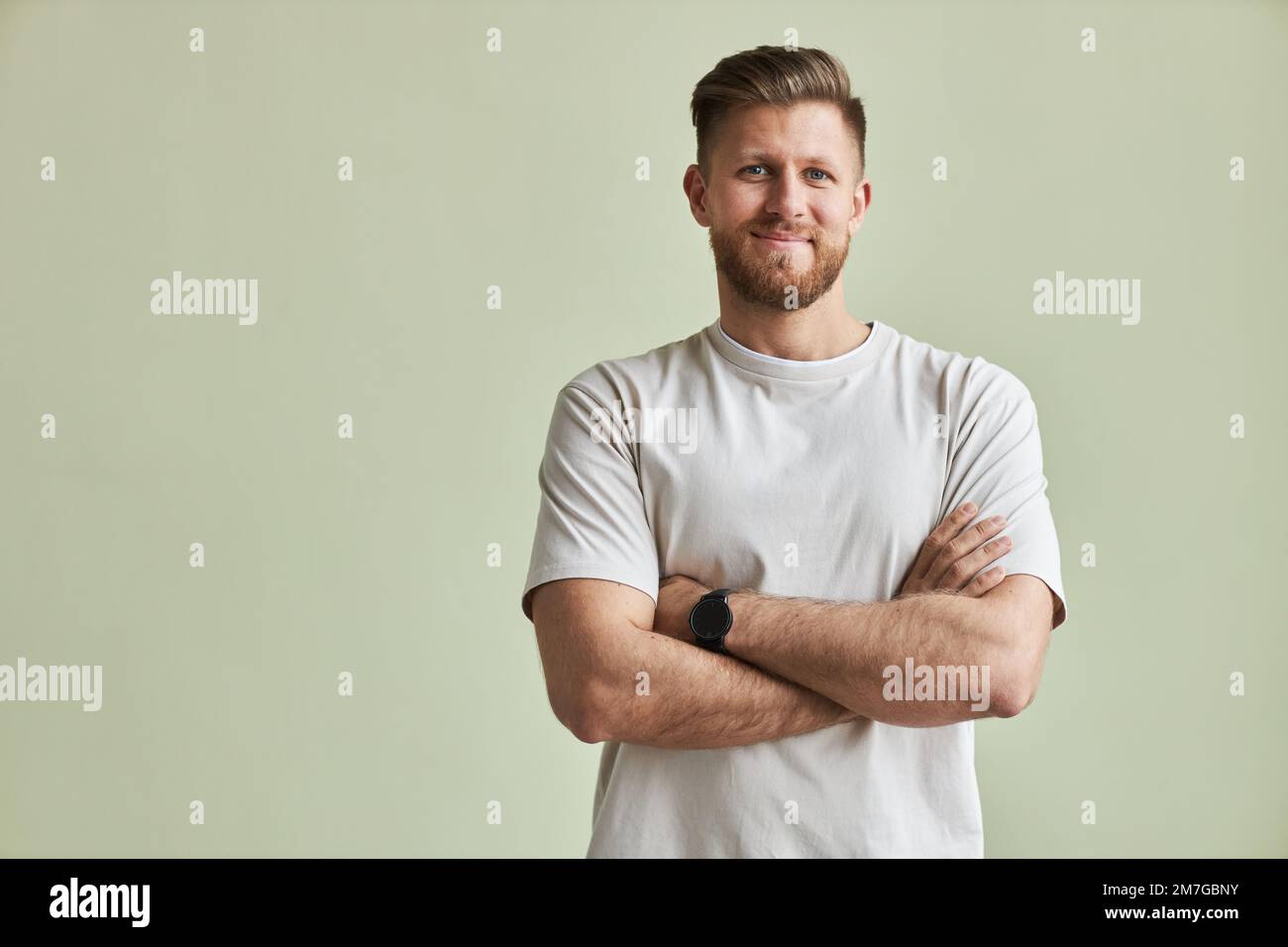 Porträt eines bärtigen jungen Mannes, der vor der Kamera lächelt, während er mit gekreuzten Armen gegen pastellgrün, minimal steht Stockfoto