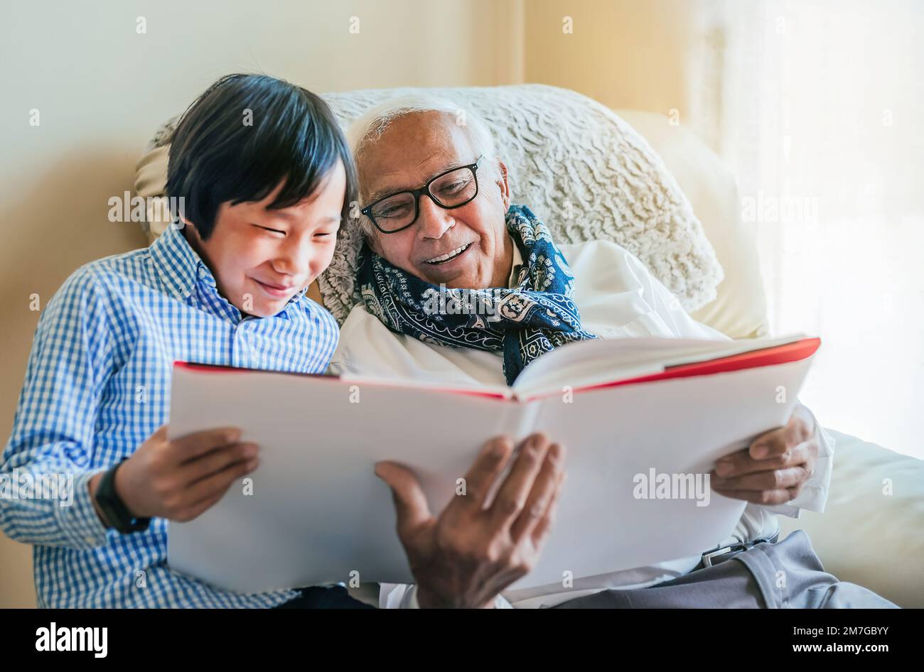 Ein weißer Großvater und sein asiatischer Enkel lesen ein Buch und amüsieren sich im Wohnzimmer. Das Licht ist weich. Konzeption und Familie. Stockfoto