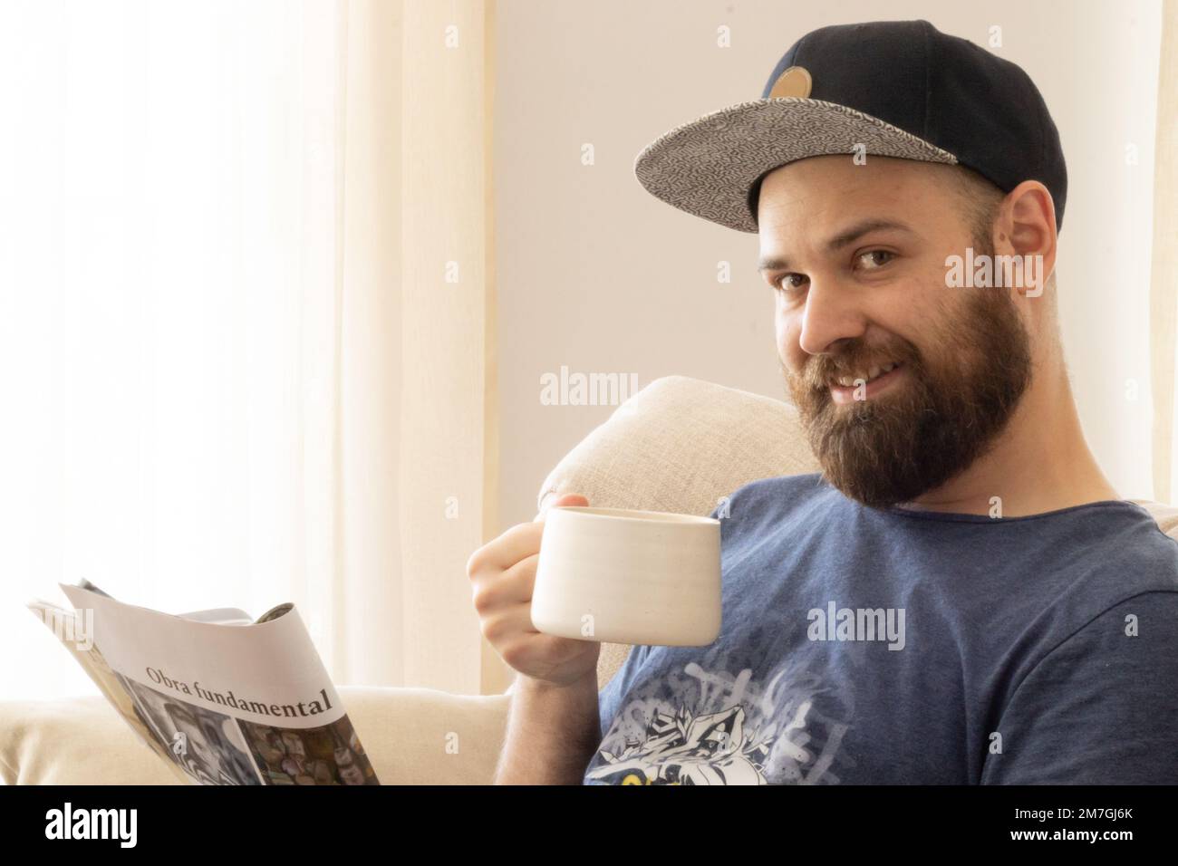 Ein weißer bärtiger Mann, der eine Tasse Kaffee genießt, während er eine Zeitschrift liest. „Obra Fundamental“ bedeutet grundlegende Arbeit. Stockfoto
