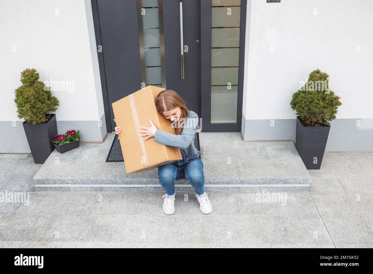 Eine weiße Frau, die auf einer Stufe vor der Haustür sitzt und eine Kiste umarmt, die gerade mit der Post kam Stockfoto