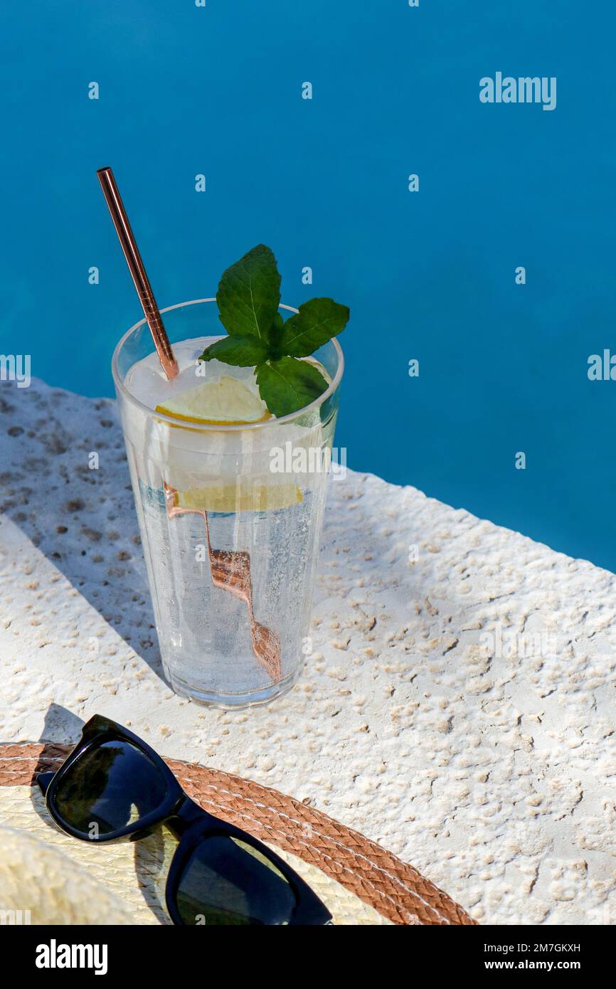 Erfrischendes Glas Wasser mit Zitrone, Strohhut und Sonnenbrille am Pool Stockfoto