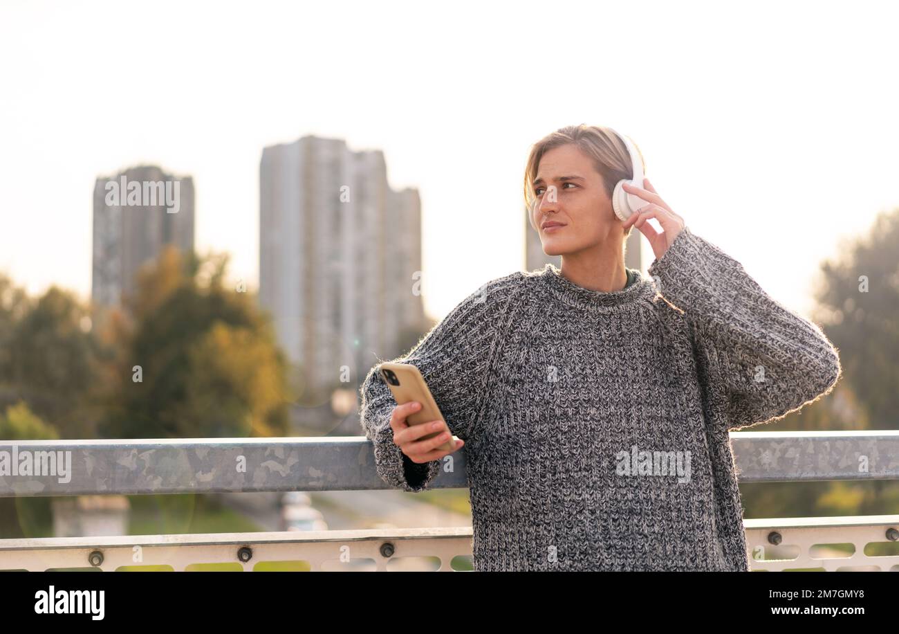 Stilvolle blonde Frau mit kurzen Haaren in Kopfhörern, die ihr Handy in der Hand hält und auf einer Brücke gegen die Stadt steht. Stockfoto