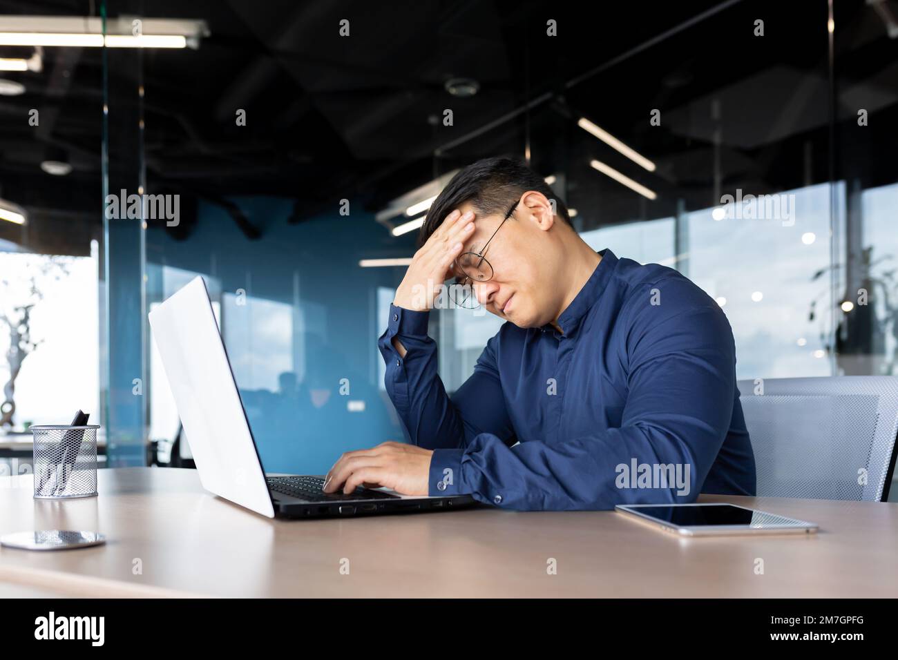Ein müder junger Asiate sitzt im Büro am Tisch, hält seinen Kopf, fühlt Kopfschmerzen, Spannung, Migräne nach einem harten Überstunden-Tag, zittert. Stockfoto