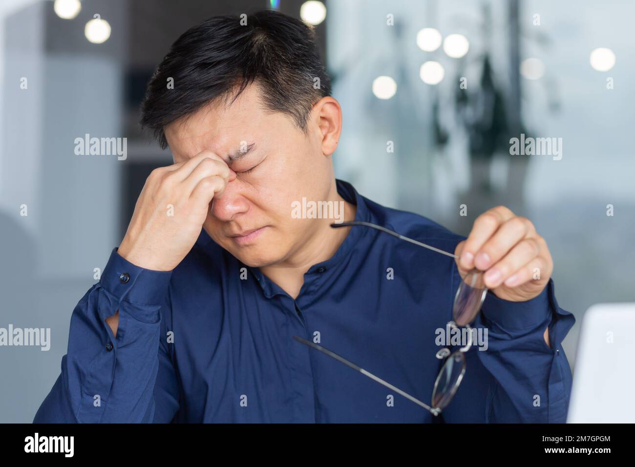 Nahaufnahme. Burnout bei der Arbeit. Ein müder junger Asiate sitzt im Büro, hält eine Brille in der Hand und schließt seine Augen mit der Hand. Ruht, spürt Spannung, Last. Braucht Ruhe, Pausen. Stockfoto