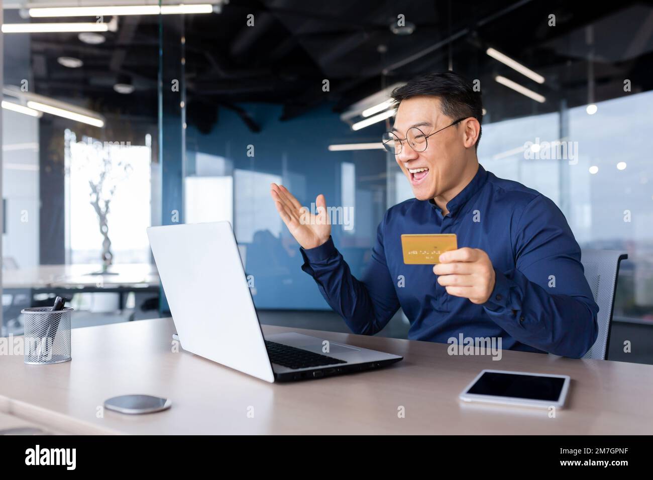 Ein glücklicher junger Asiate sitzt mit einem Laptop im Büro und hält eine Kreditkarte in der Hand. Online-Zahlung, Online-Shopping, Geldtransfer, Kontoüberprüfung. Stockfoto