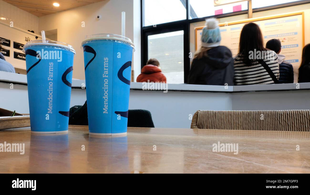 Zwei Getränketassen auf einem Tisch, während Gäste im Hintergrund die Speisekarte und die Speisemöglichkeiten eines Sandwichgeschäfts der Mendocino Farms in San Jose, Kalifornien, betrachten Stockfoto