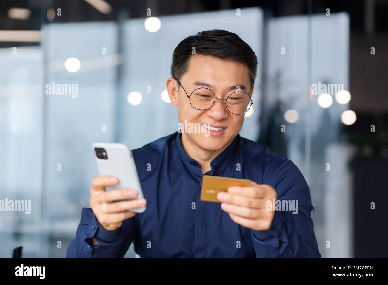 Nahaufnahme. Ein glücklicher junger Asiate sitzt im Büro am Tisch und hält eine Kreditkarte und ein Telefon in der Hand. Online-Zahlung, Online-Shopping, Geldtransfer, Kontoüberprüfung. Stockfoto