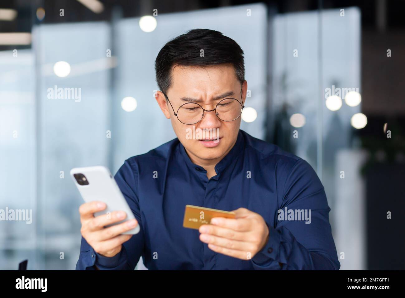 Ein besorgter junger Mann sitzt im Büro und hält eine Kreditkarte und ein Telefon in der Hand. Erfolgloses Online-Shopping, Transaktion, Geldmangel auf dem Konto, gesperrtes Konto. Bankrott. Stockfoto