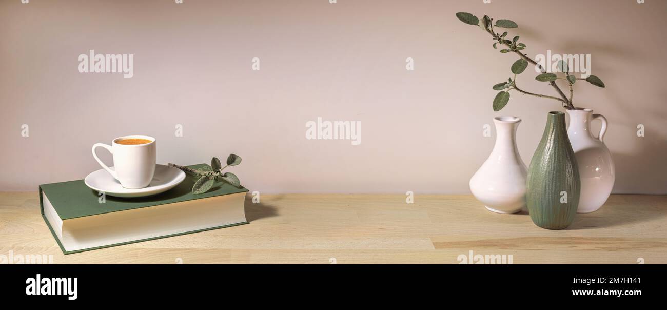 Kaffeetasse auf einem grauen grünen Buch und eine Gruppe von Vasen mit einem Salbeiblatt Ast auf einem Holztisch vor einer hellen Wand, Panoramaformat, Inneneinrichtung Stockfoto