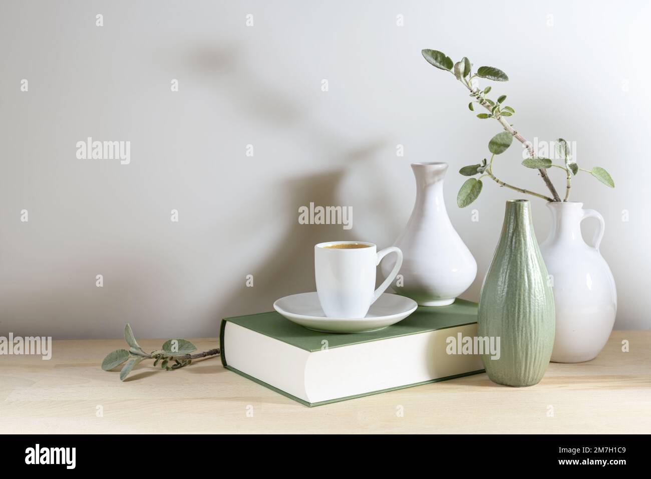 Noch immer leben von drei Vasen mit einem Salbeiblatt-Zweig, einem grauen grünen Buch und einer Kaffeetasse auf einem Holztisch oder Tisch vor einer hellgrauen Wand, ruhiges Zuhause Stockfoto