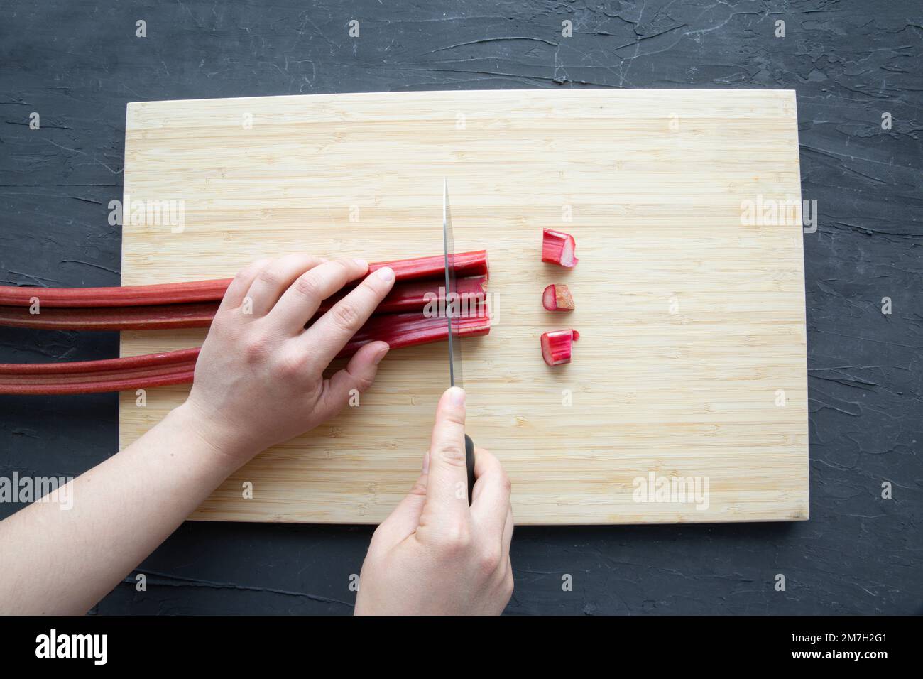 Frischer Rhabarber auf Holzspanplatte auf schwarzem Hintergrund. Innengewinde hält Messer und Häckseln, Draufsicht. Stockfoto