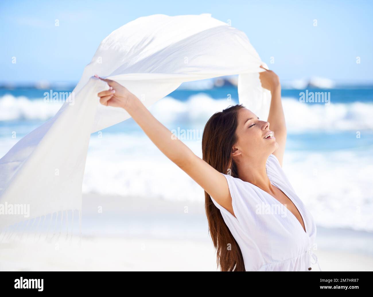 Schönheit in der Brise. Eine wunderschöne junge Frau am Strand mit einem weißen Schal, der im Wind weht. Stockfoto