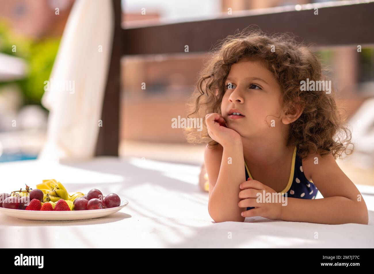 Süßes, lockiges Mädchen, das mit einem Teller mit Früchten neben ihr liegt Stockfoto