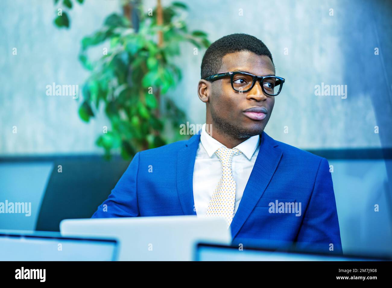 Afroamerikanischer Unternehmer in blauem Anzug und stilvoller Brille, der während der Arbeit in einem modernen Büro auf dem Laptop wegsieht Stockfoto