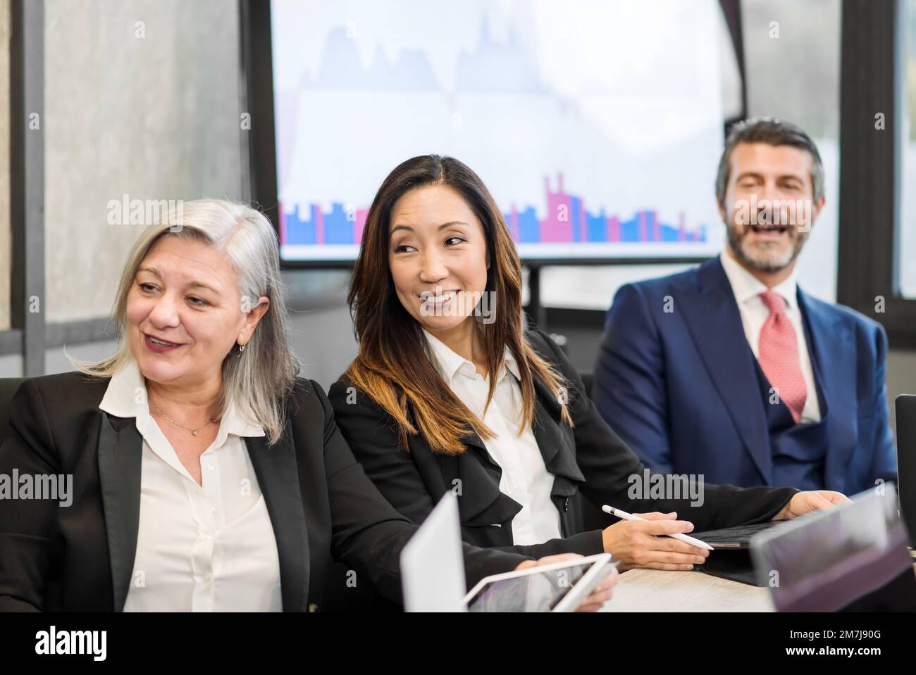 Multiethnische Kollegen in formellen Kleidern lächeln und schauen weg, während sie während einer Besprechung an einem Tisch in einem modernen Sitzungssaal sitzen Stockfoto